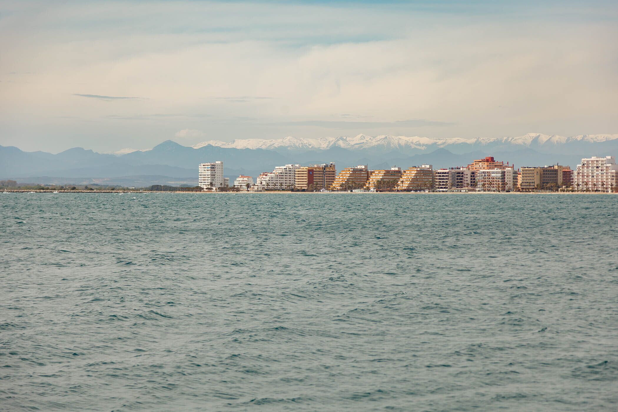 A waterfront cityscape with a row of multi-story buildings, a calm sea, and snow-capped mountains in the background.