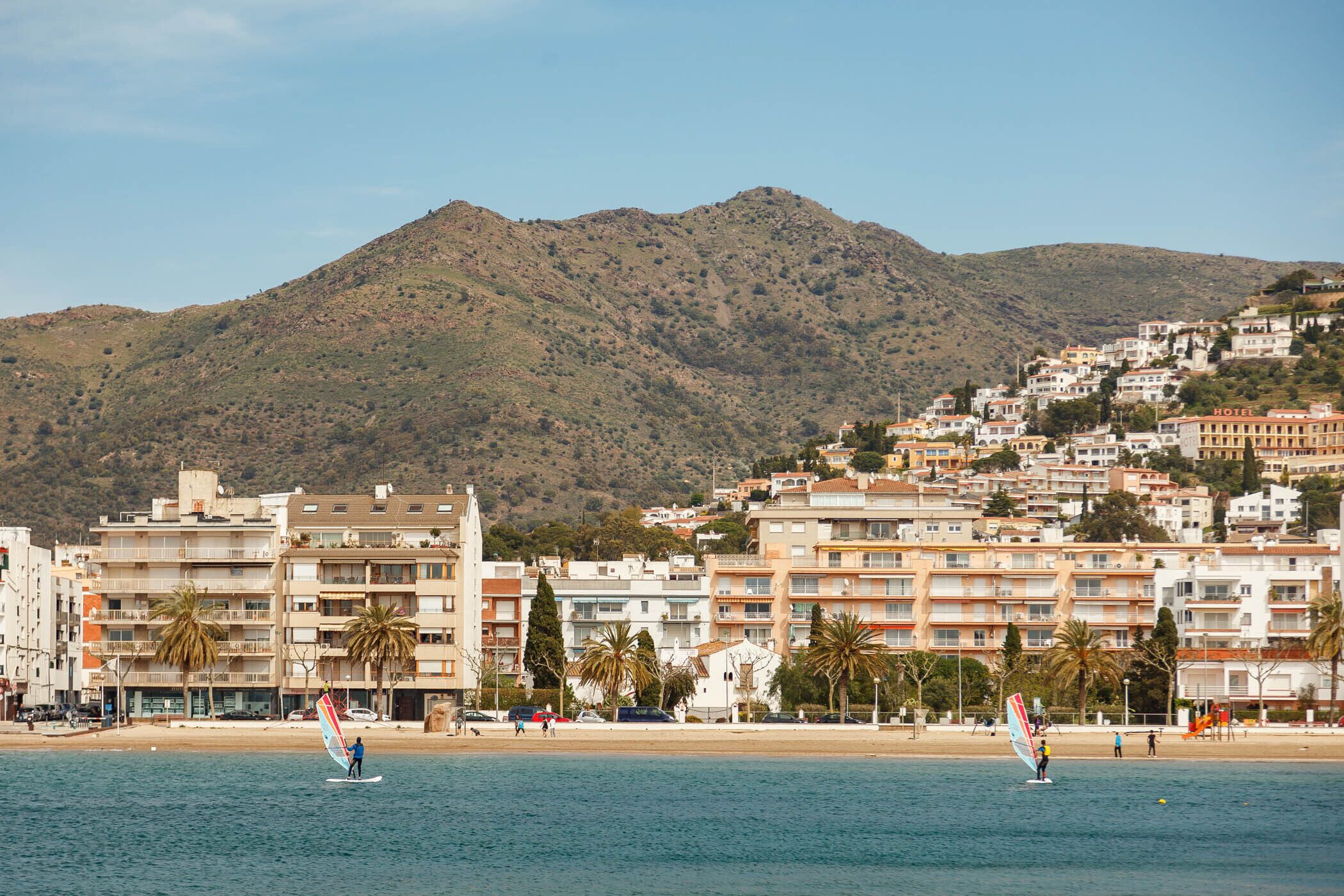 Ein Küstenhotel mit mehreren Etagen, Balkonen und Blick auf das Meer sowie einen Berg im Hintergrund.