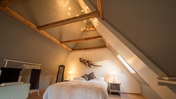 A bedroom with a sloped ceiling, wooden beams, a bed with a white bedspread, a nightstand with a lamp, and a wall-mounted mirror.