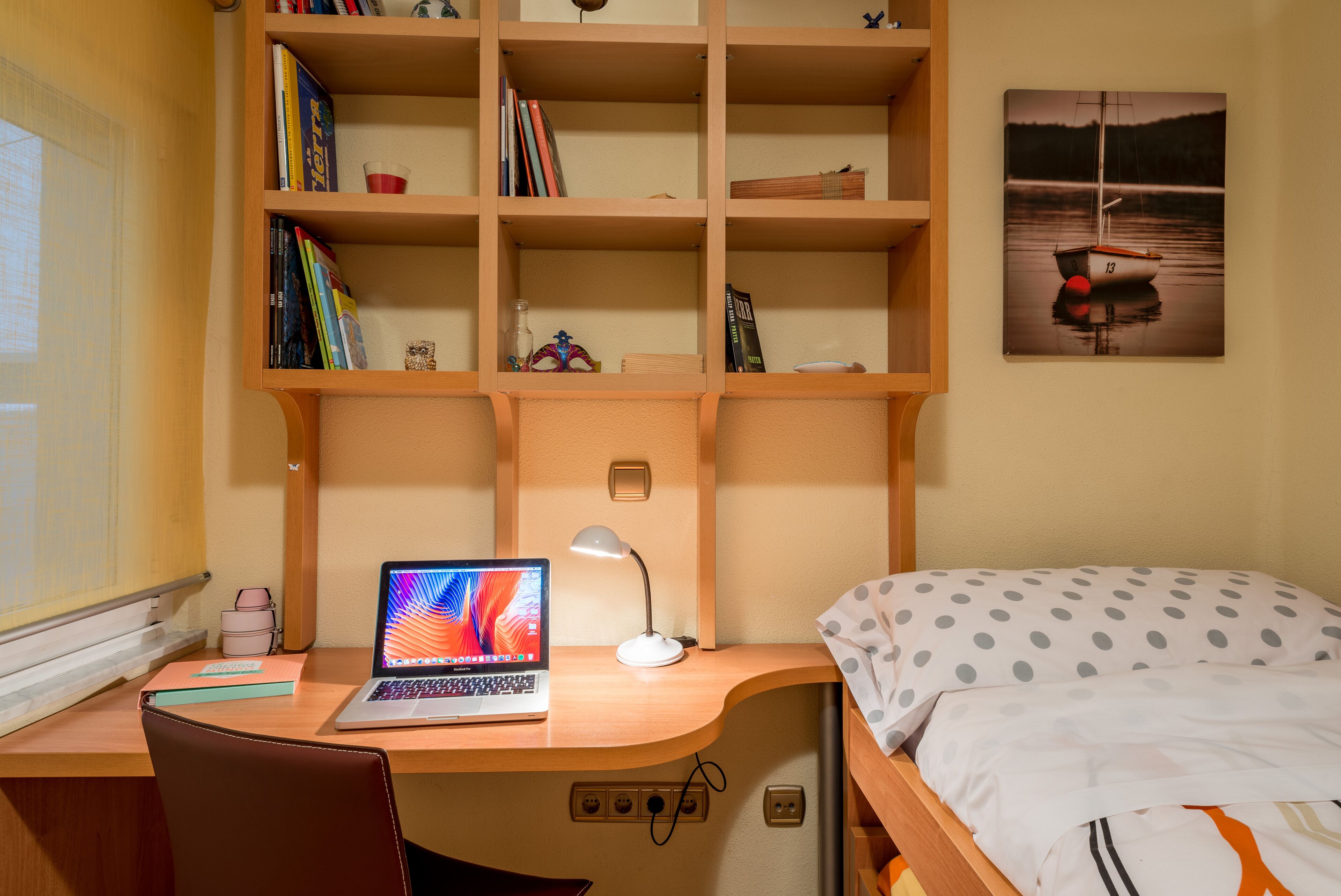 Una camera da letto con una scrivania in legno, un laptop, una lampada, un letto con copriletto a pois e una libreria con libri e oggetti decorativi.