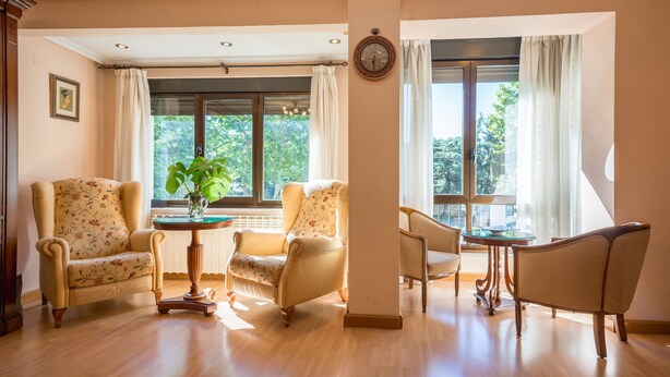 A room with a wooden floor, two armchairs, a round table, and a window with curtains.