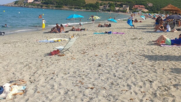 Ein Strand mit Sonnenschirmen, Personen, die sich sonnen, und eine Hügelkette mit Gebäuden im Hintergrund.