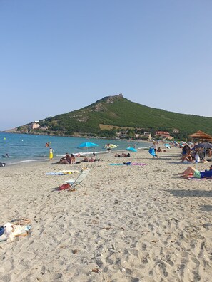 A beach with umbrellas, people sunbathing, and a hill with buildings in the background.