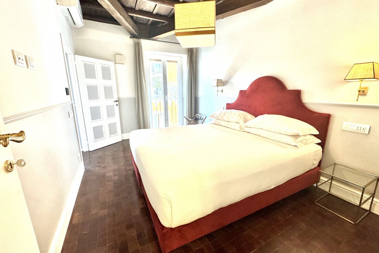 A hotel room with a bed, a door, a window, and a wall-mounted lamp.