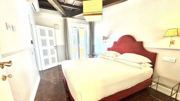 A hotel room with a bed, a door, a window, and a wall-mounted lamp.
