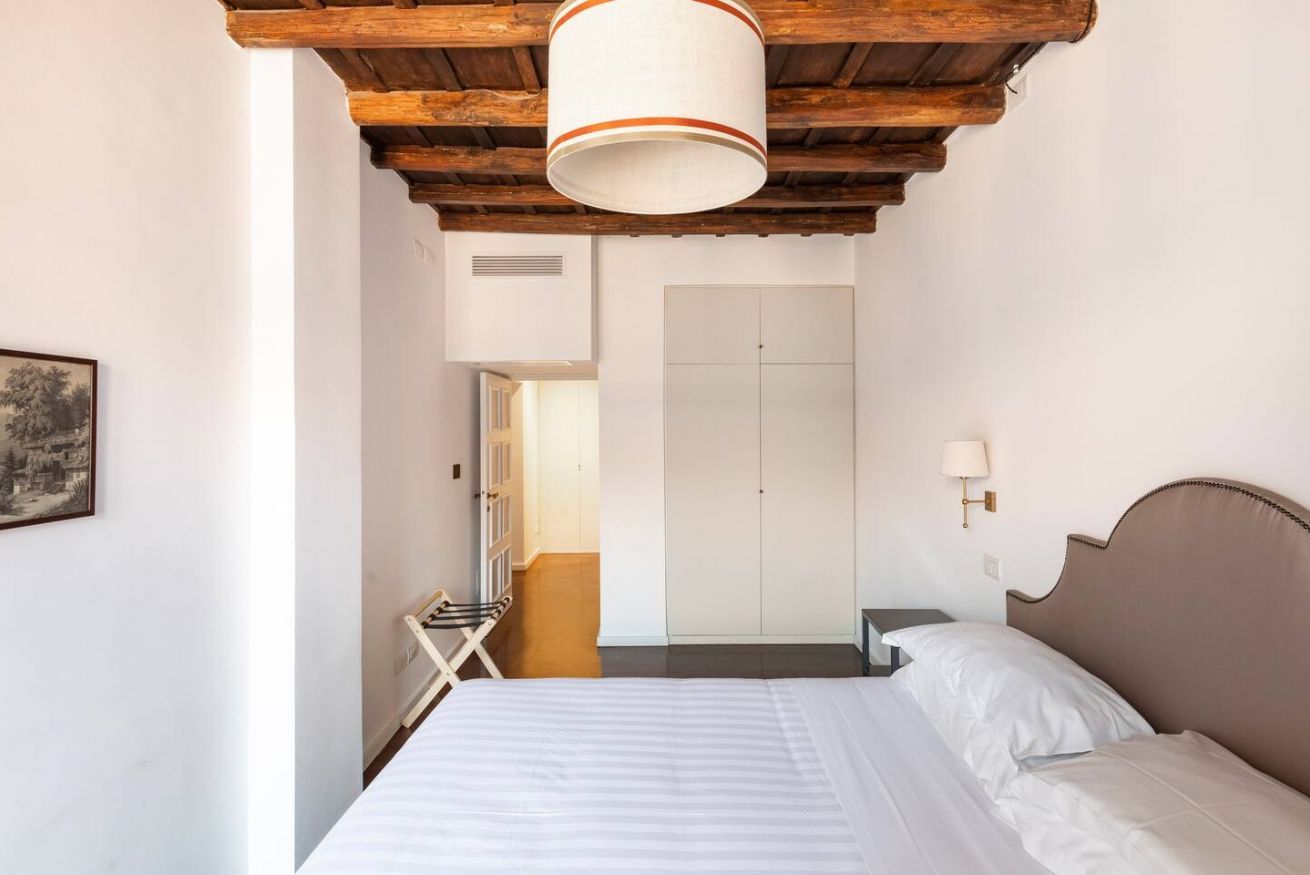Een slaapkamer met een houten plafond, een bed met witte lakens, een nachtkastje en een wandlamp.