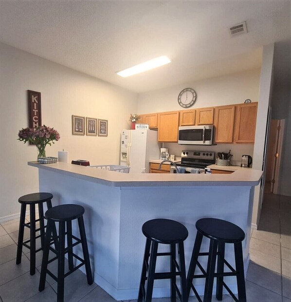 A kitchen with wooden cabinets, a microwave, a refrigerator, and a clock on the wall. There are three black stools at the island.