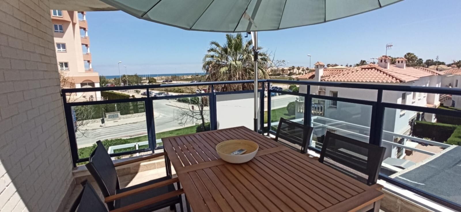 A balcony with a wooden table and chairs, a bowl, and a view of a street and buildings.