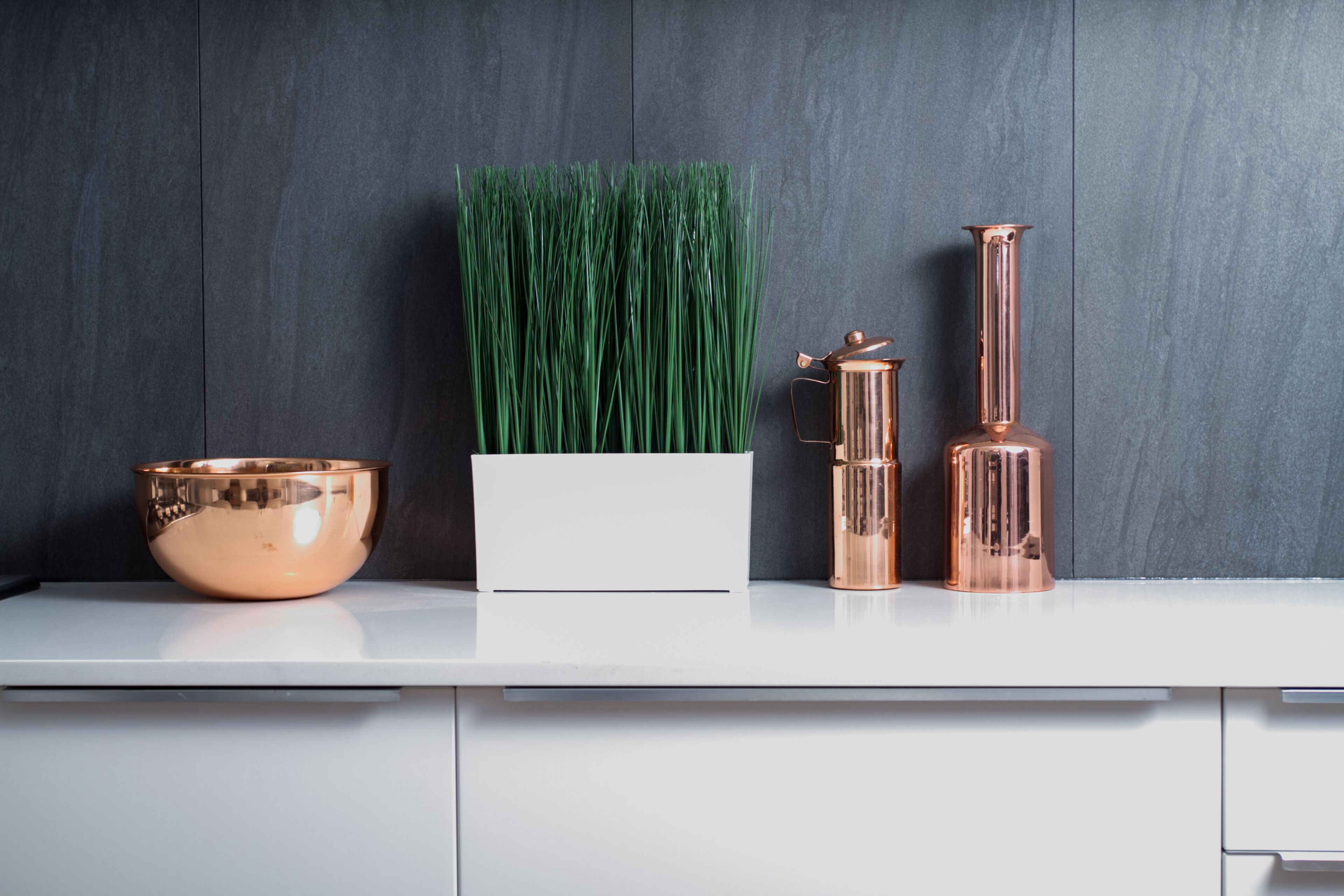 Un plan de travail blanc surmonté d’un saladier en cuivre, un pot de fleurs blanc contenant de l’herbe verte et deux vases en cuivre.