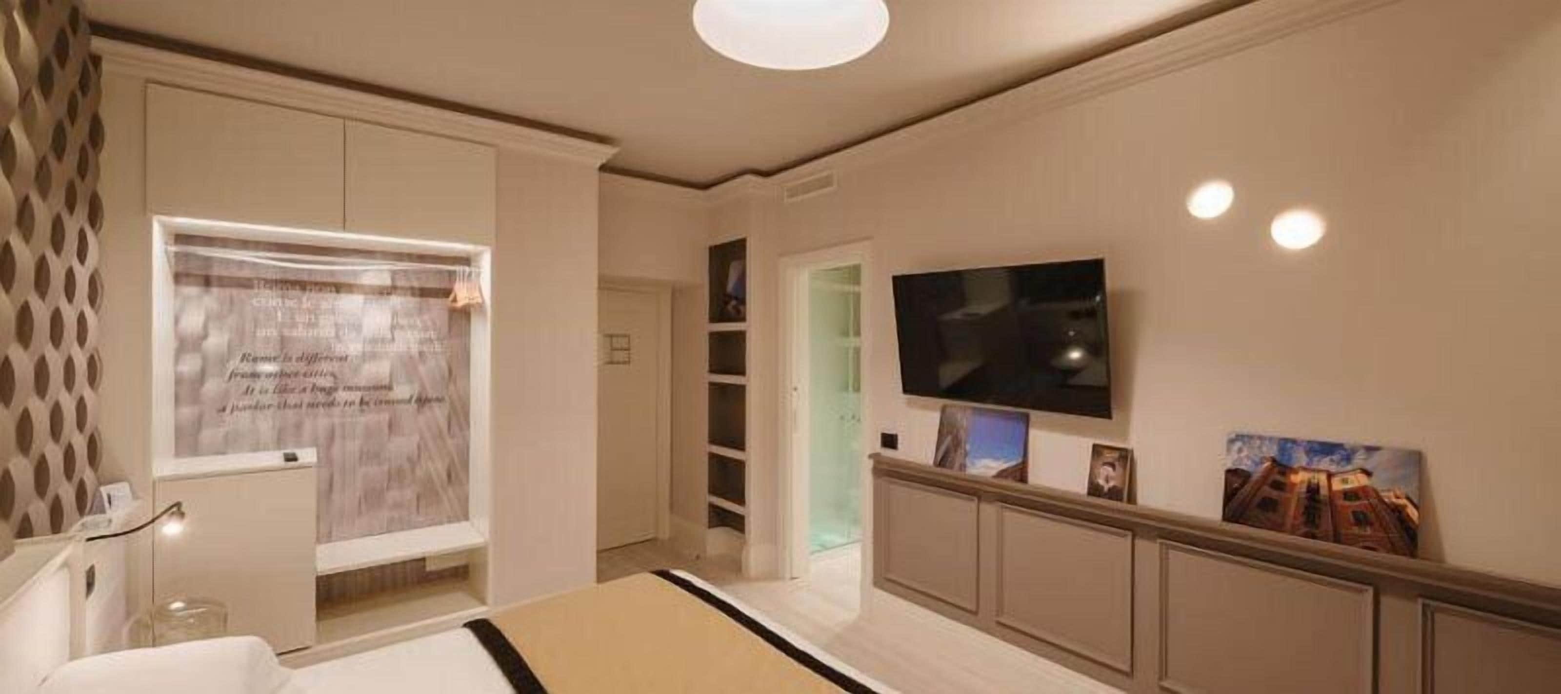 Een moderne hotelkamer met een grote flatscreen-televisie aan de muur, een bed met hoofdbord, een klein bureau en een raam met decoratieve gordijnen.