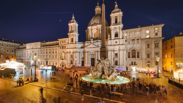 Una piazza storica con una fontana imponente, circondata da architettura classica e affollata di persone.