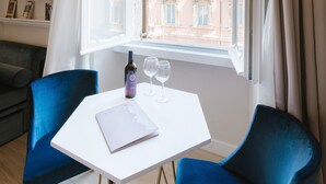 Una stanza con un tavolo bianco, due sedie blu, una bottiglia di vino e due bicchieri da vino. Una finestra con tende aperte mostra un edificio con una bandiera.