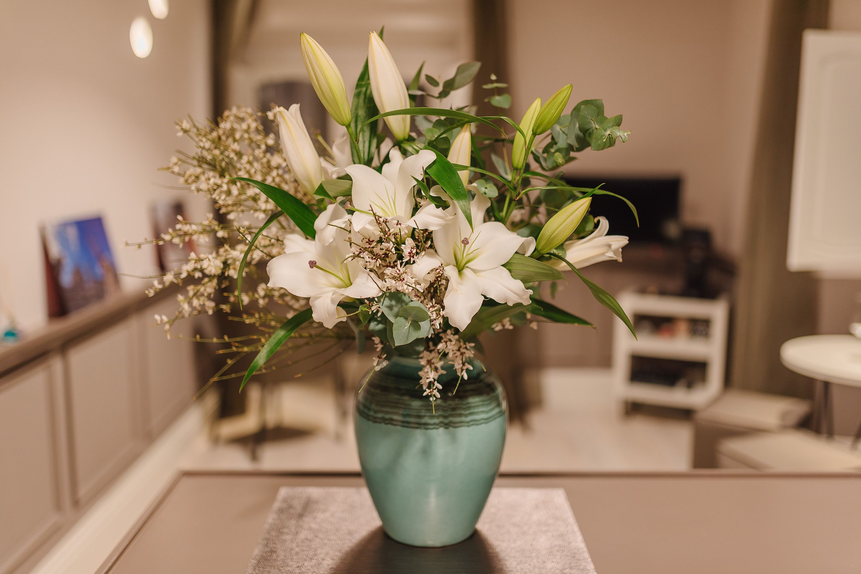 Un florero azul aguamarina con flores blancas y hojas verdes sobre una mesa en una habitación con una paleta de colores neutros y decoración moderna.