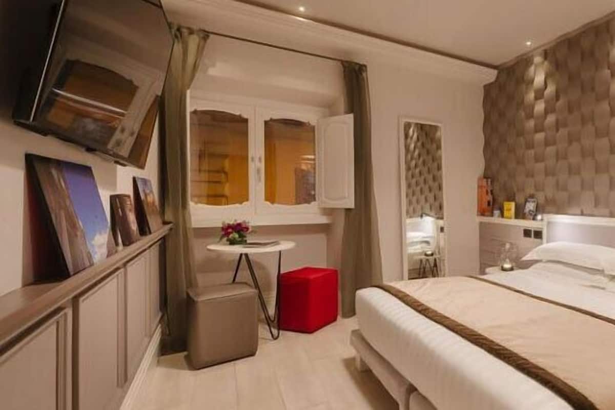 Une chambre d’hôtel moderne avec un grand lit, une petite table ronde, un pouf rouge et un téléviseur fixé au mur.
