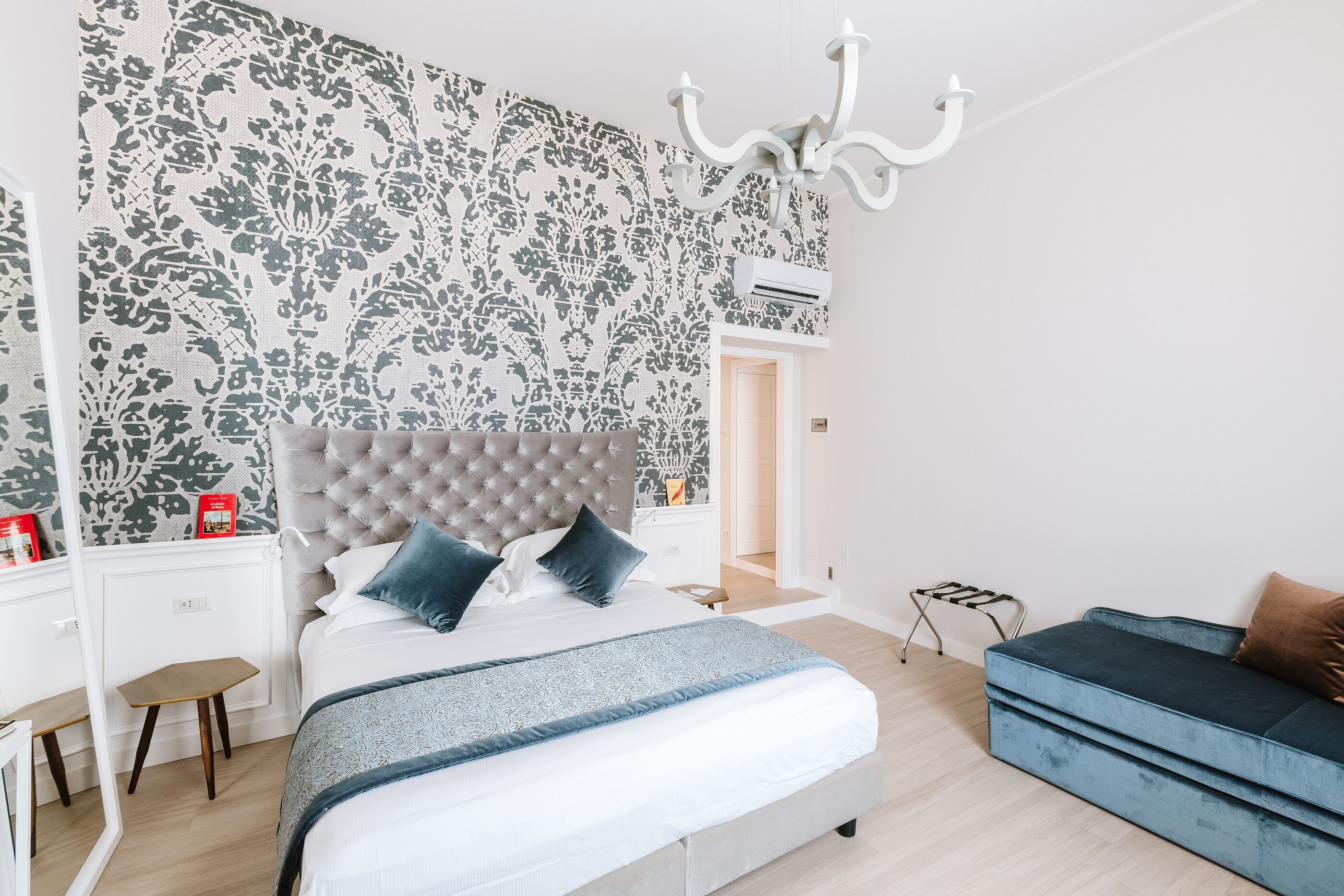 Un dormitorio con cabecera capitonada, cama con sábanas blancas y almohadas azules, sofá azul, un taburete de madera pequeño y un papel tapiz estampado.