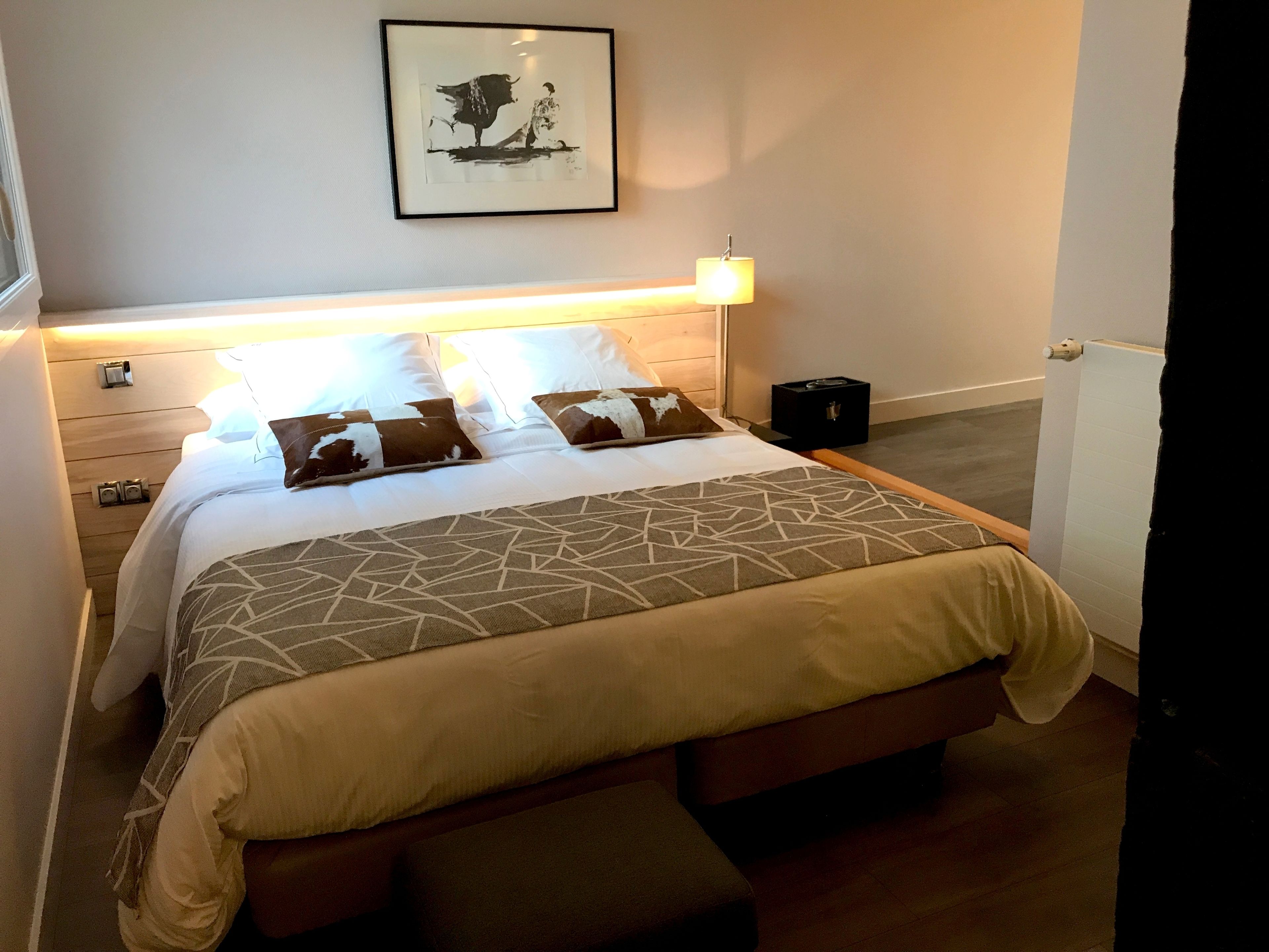 A neatly made bed with a geometric-patterned bedspread, a framed artwork above it, and a bedside lamp.