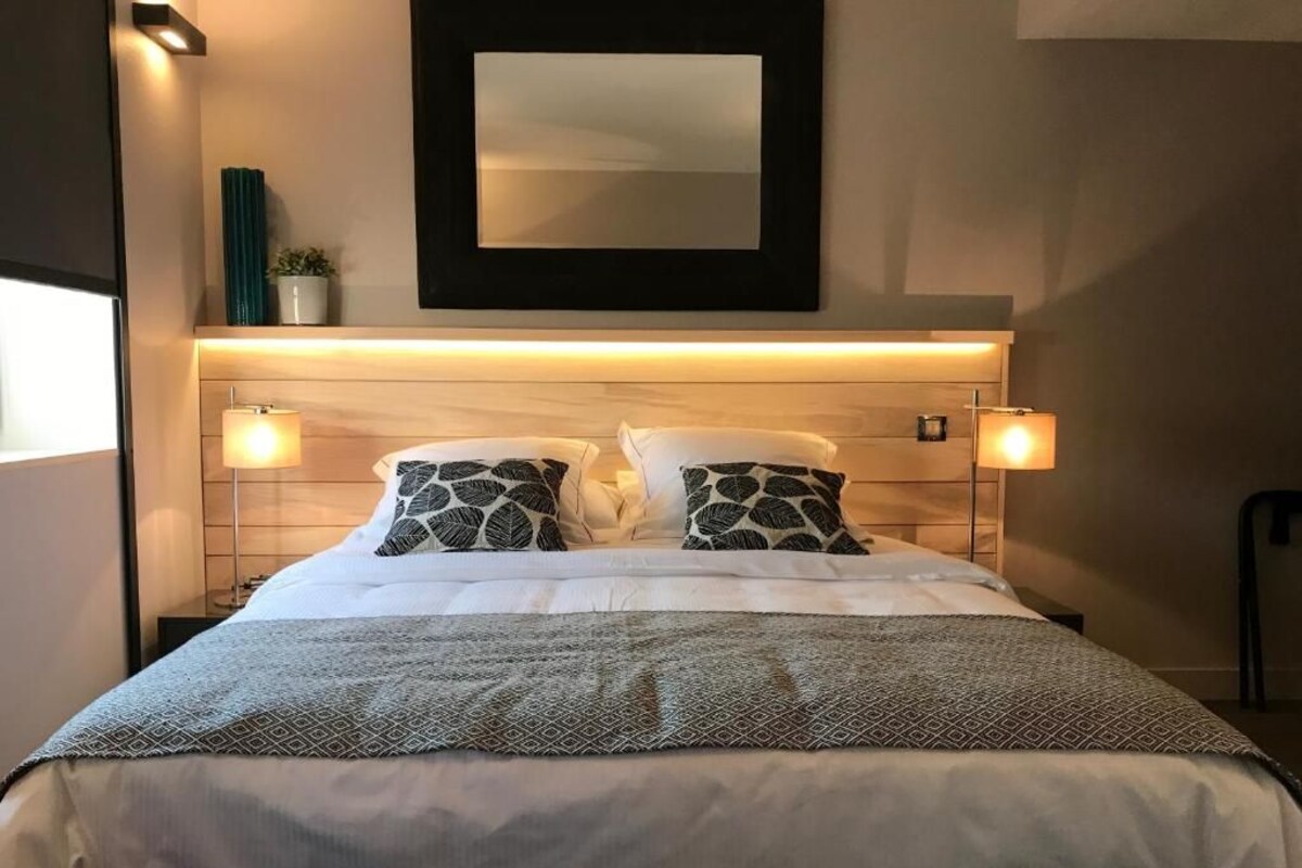 Un lit bien fait, agrémenté d’oreillers à motifs, une table de chevet avec une lampe et un miroir fixé au mur.