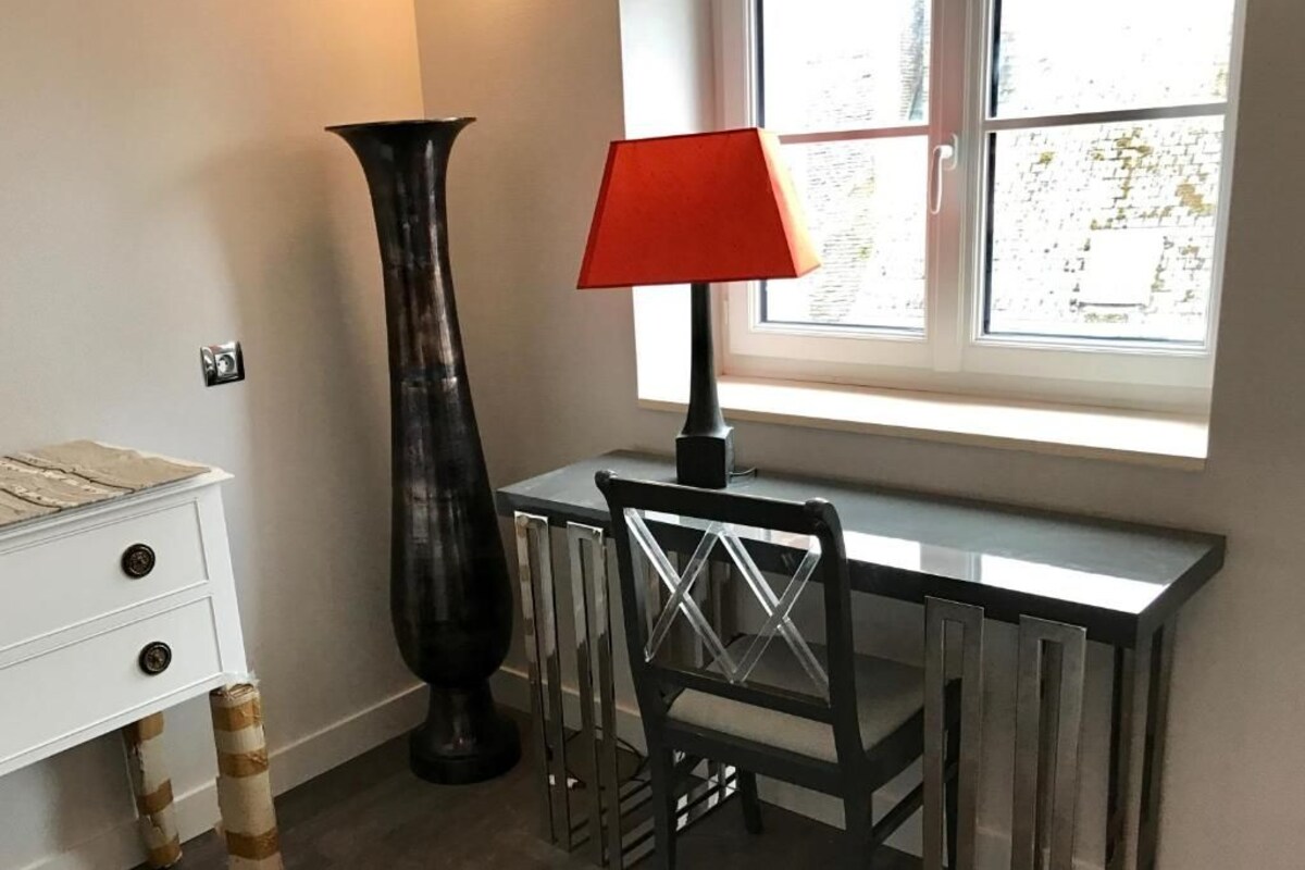 Une pièce avec une commode blanche, un grand vase noir, une chaise noire avec un dossier en verre et une lampe rouge posée sur une table noire près d’une fenêtre.