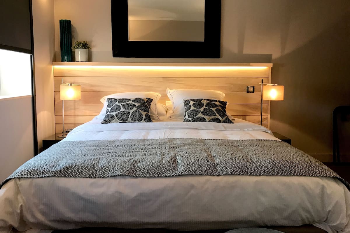 Un lit bien fait, agrémenté d’oreillers à motifs, une table de chevet avec une lampe et un miroir fixé au mur.