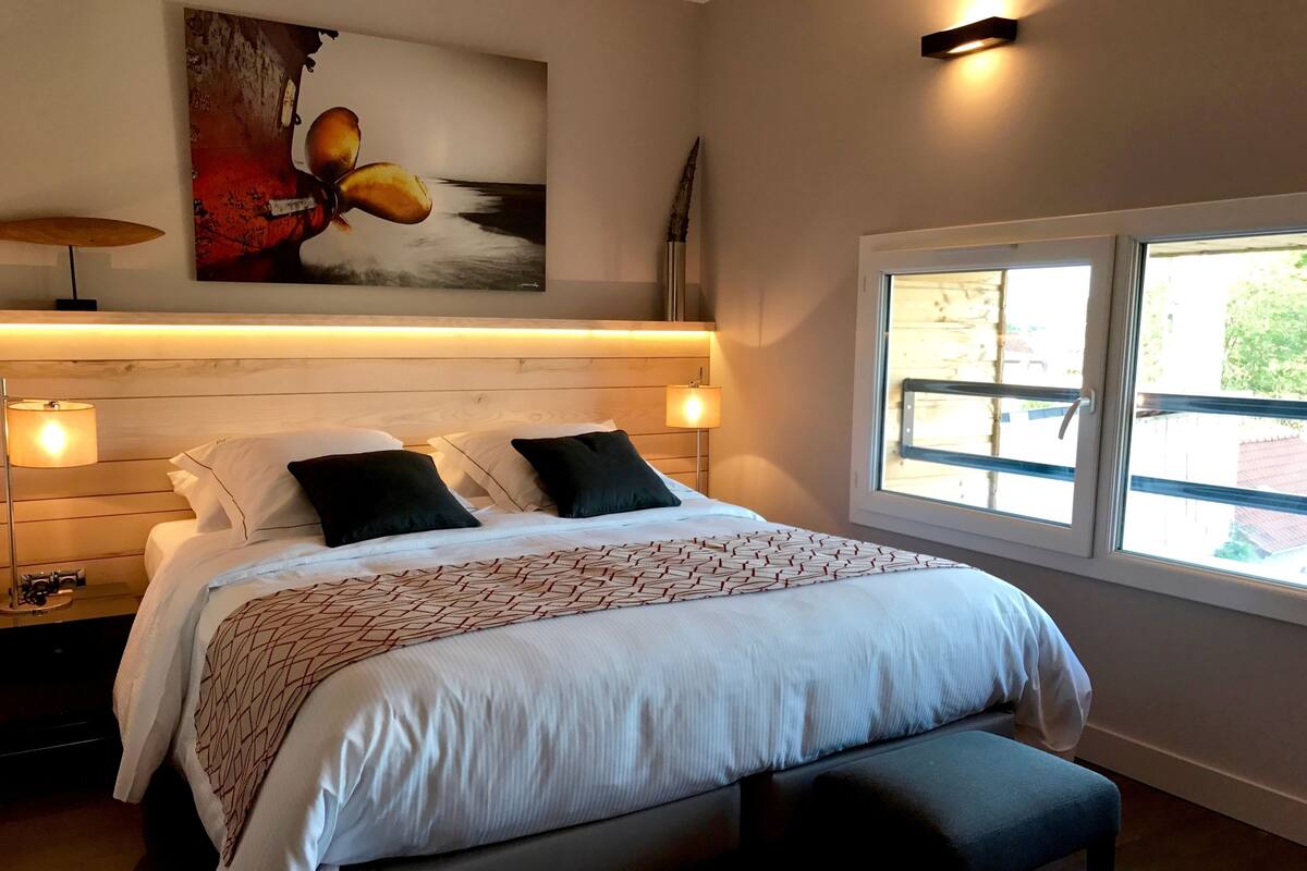 Une chambre avec un grand lit, deux lampes de chevet, une table de chevet, un banc, une fenêtre donnant sur un beau paysage et un tableau accroché au mur.