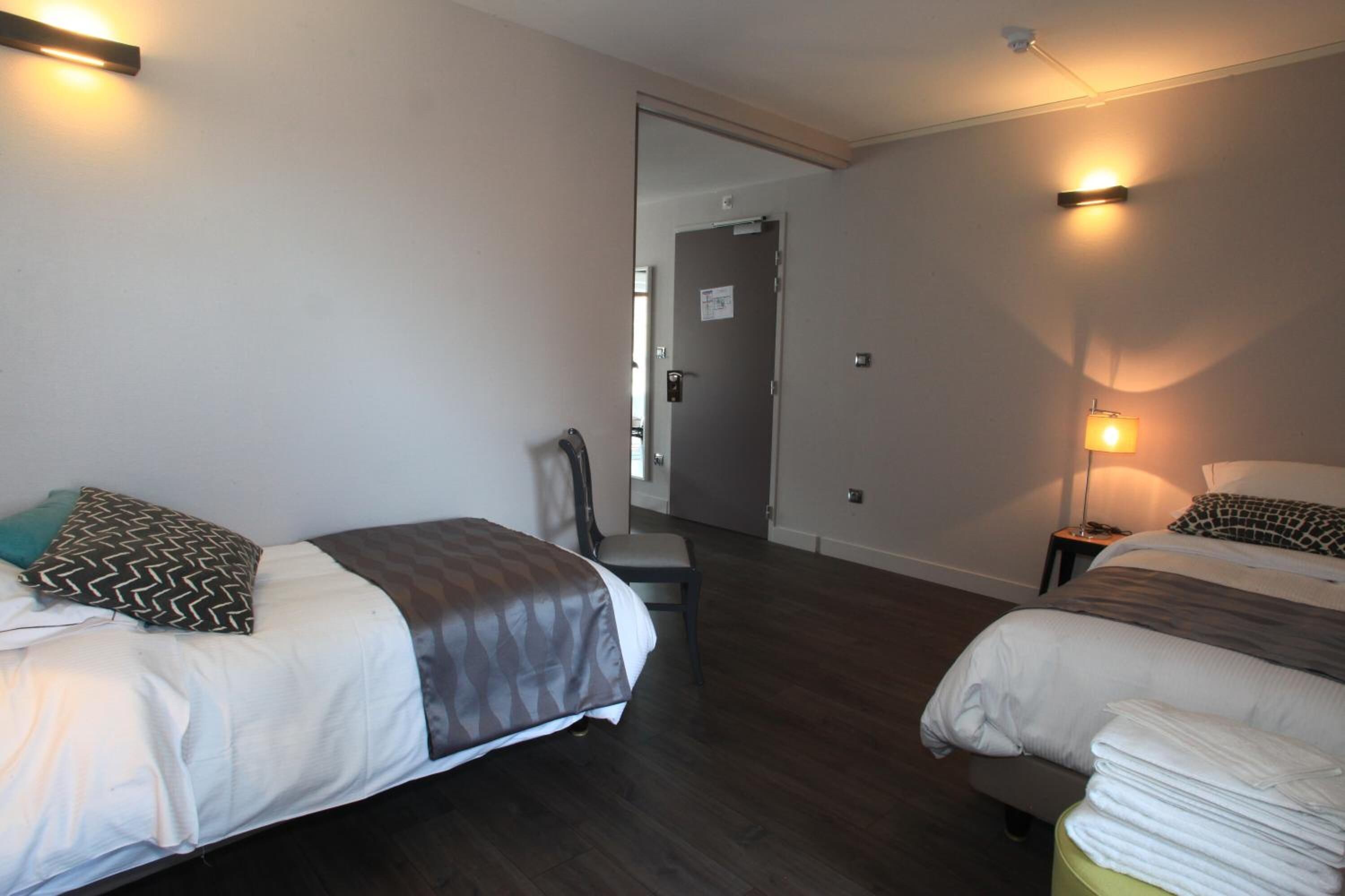A hotel room with two beds, a chair, a small table, and a lamp.