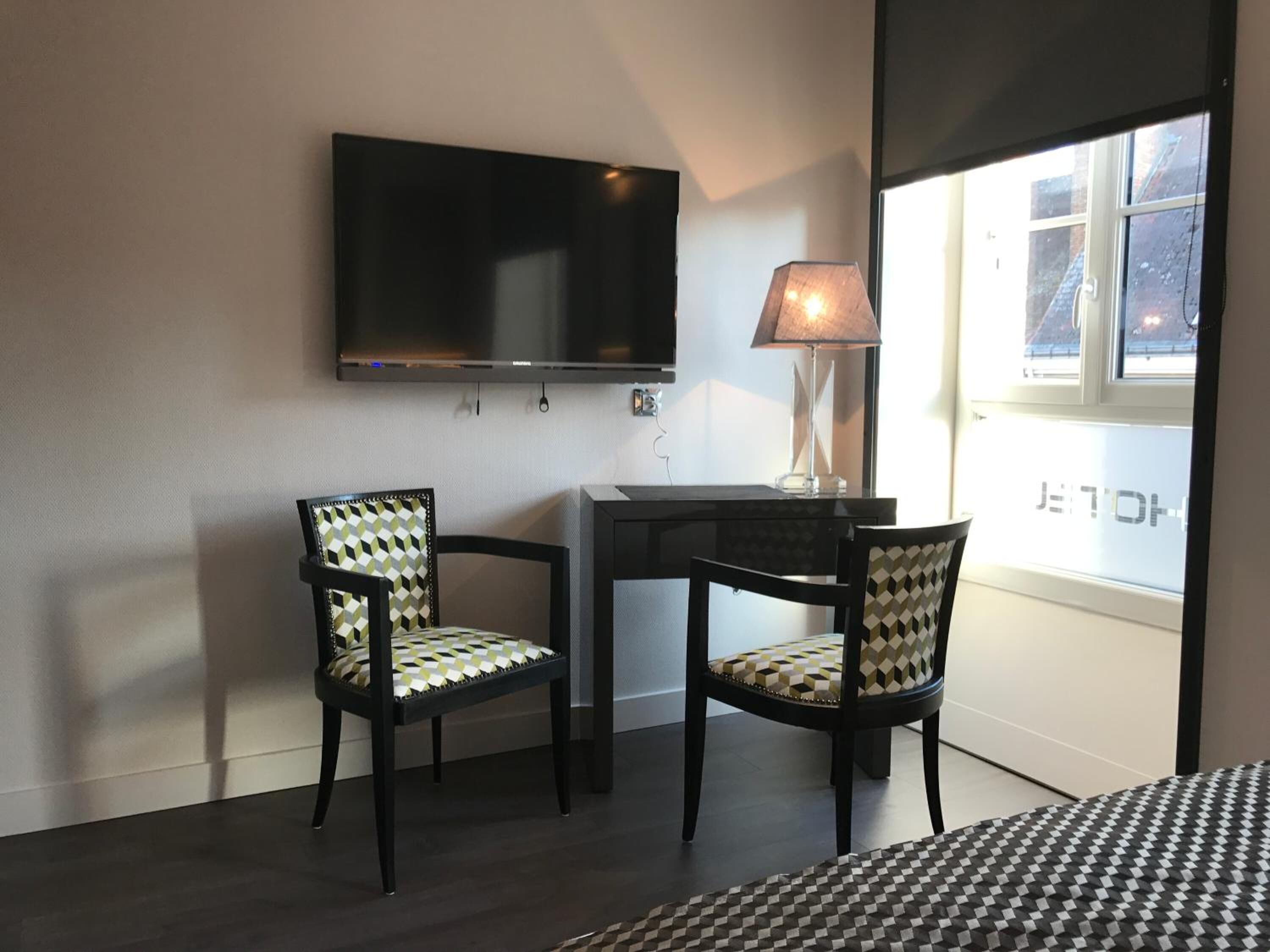 A small room with a TV mounted on the wall, two chairs with patterned cushions, a desk with a lamp, and a window with a view of the outside.