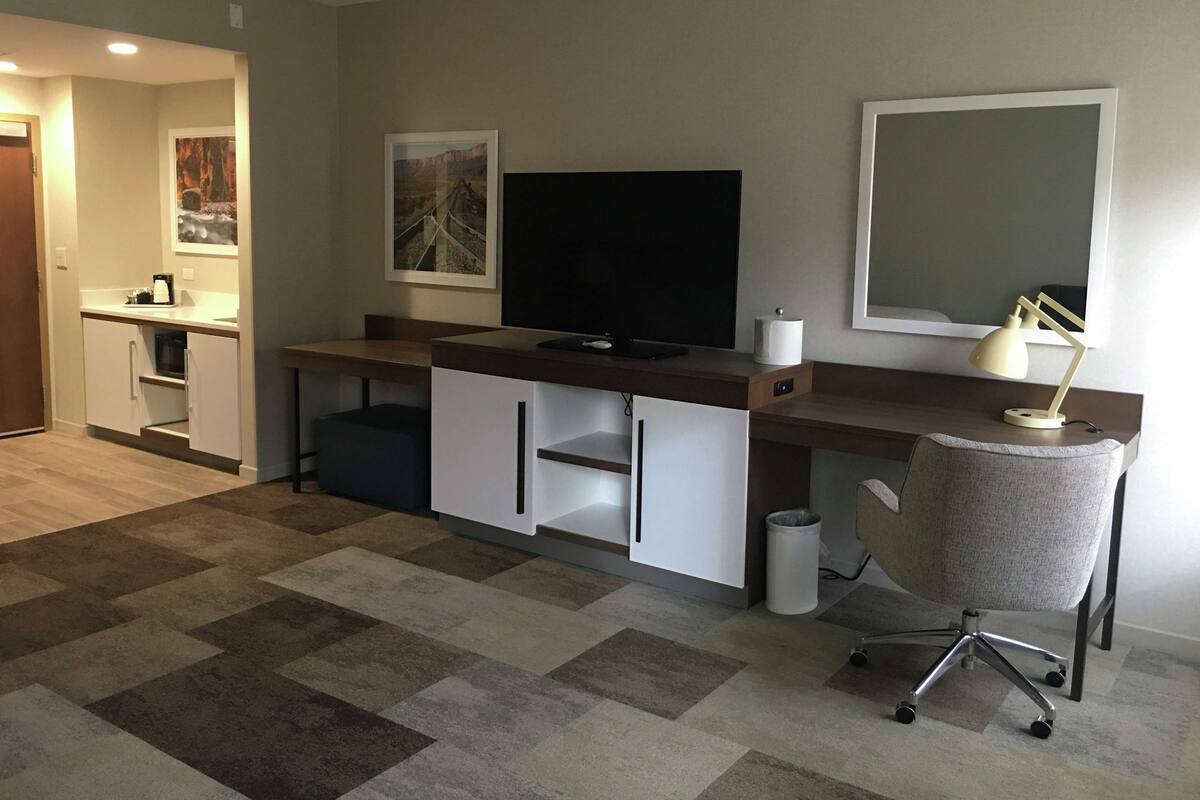 Una sala de estar moderna con un televisor de pantalla plana, un escritorio con lámpara, una silla y una pequeña zona de kitchenette.