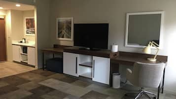 A modern living room with a flat-screen TV, a desk with a lamp, a chair, and a small kitchenette area.