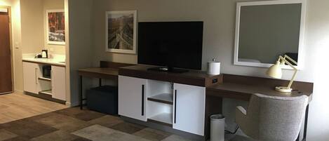 Una sala de estar moderna con un televisor de pantalla plana, un escritorio con lámpara, una silla y una pequeña zona de kitchenette.