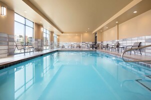 Una piscina interior con agua azul cristalina, rodeada de sillas y mesas, ventanas amplias y un muro de azulejos.