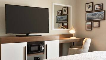 A hotel room with a bed, a TV, a desk with a lamp, a chair, a microwave, and framed pictures on the wall.