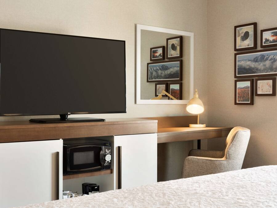 A hotel room with a bed, a TV, a desk with a lamp, a chair, a microwave, and framed pictures on the wall.