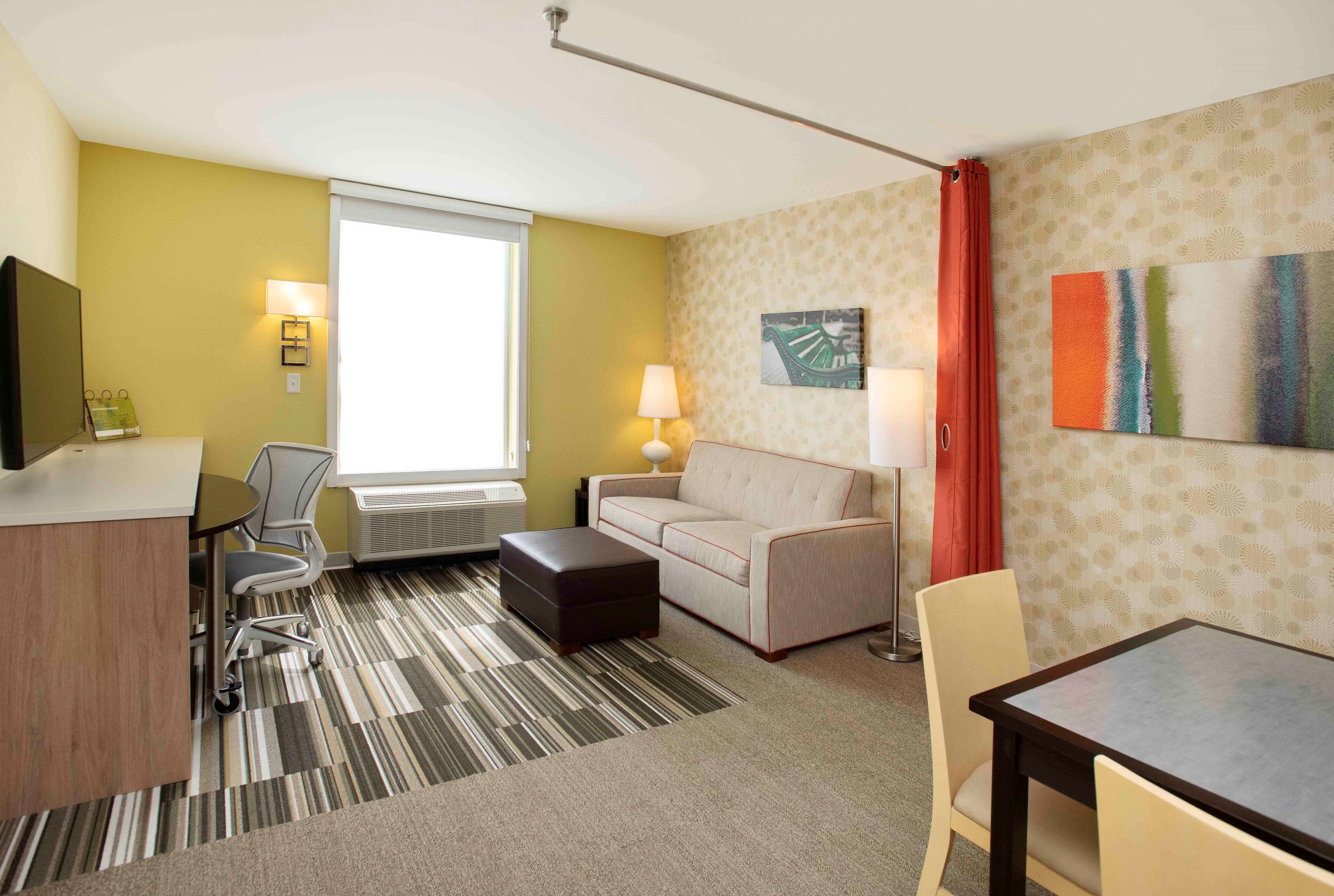 A hotel room with a sofa, a dining table, a desk, and a chair. There is a window with curtains, a lamp, and a painting on the wall.