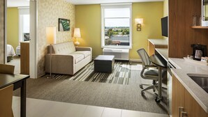 A hotel room with a sofa, a desk, a chair, a window with a view, and a kitchenette. - Home2 Suites by Hilton Mishawaka South Bend, IN (Mishawaka)