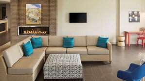 A modern living room with a sectional sofa, a fireplace, a flat-screen TV, and a dining area with red chairs. - Home2 Suites by Hilton Mishawaka South Bend, IN (Mishawaka)