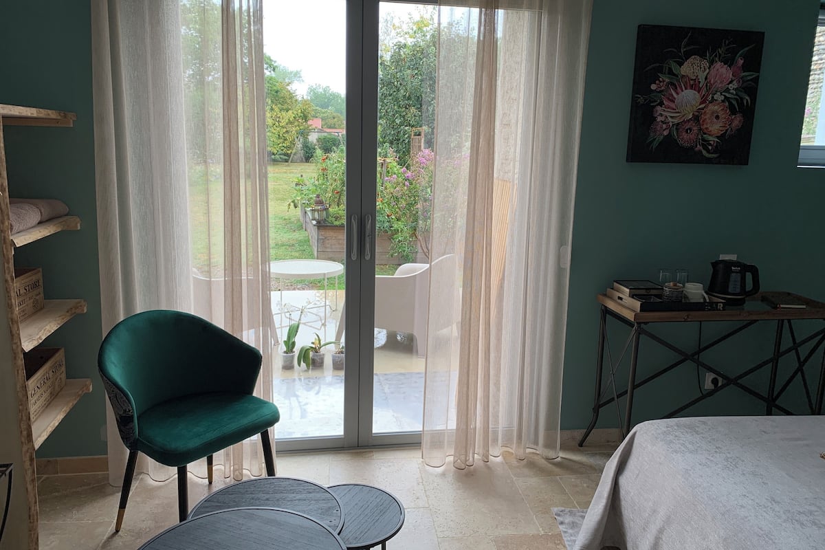 Una habitación con una silla verde, una mesita, una cama, un cuadro y vista a un jardín a través de un ventanal.