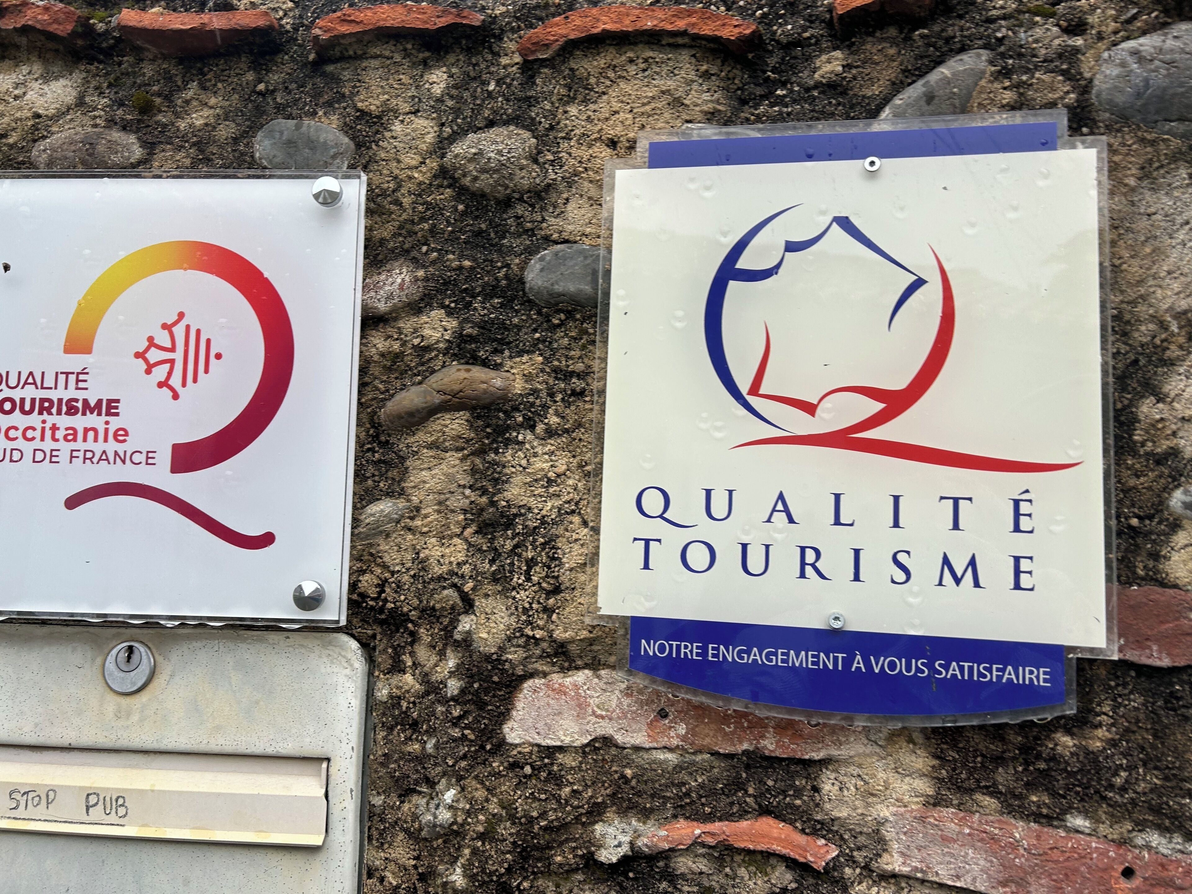 Dos señales colocadas en una pared de piedra, una con un logotipo y texto en francés, y la otra con un logotipo y texto en inglés, ambas relacionadas con la calidad turística.
