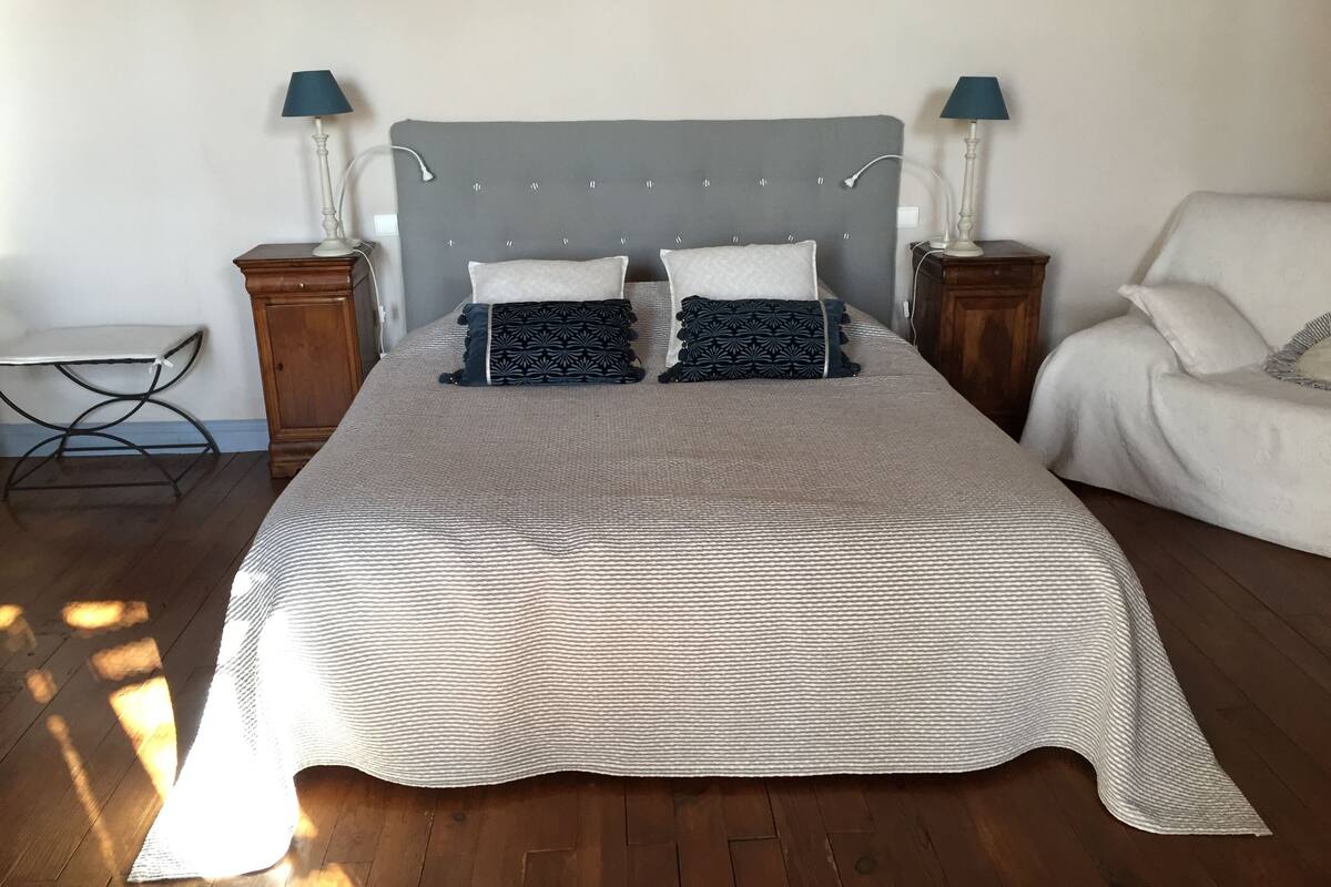 Una cama bien hecha con cabecera gris, dos mesitas de noche con lámparas, una pequeña mesa y una silla en una habitación con suelo de madera.