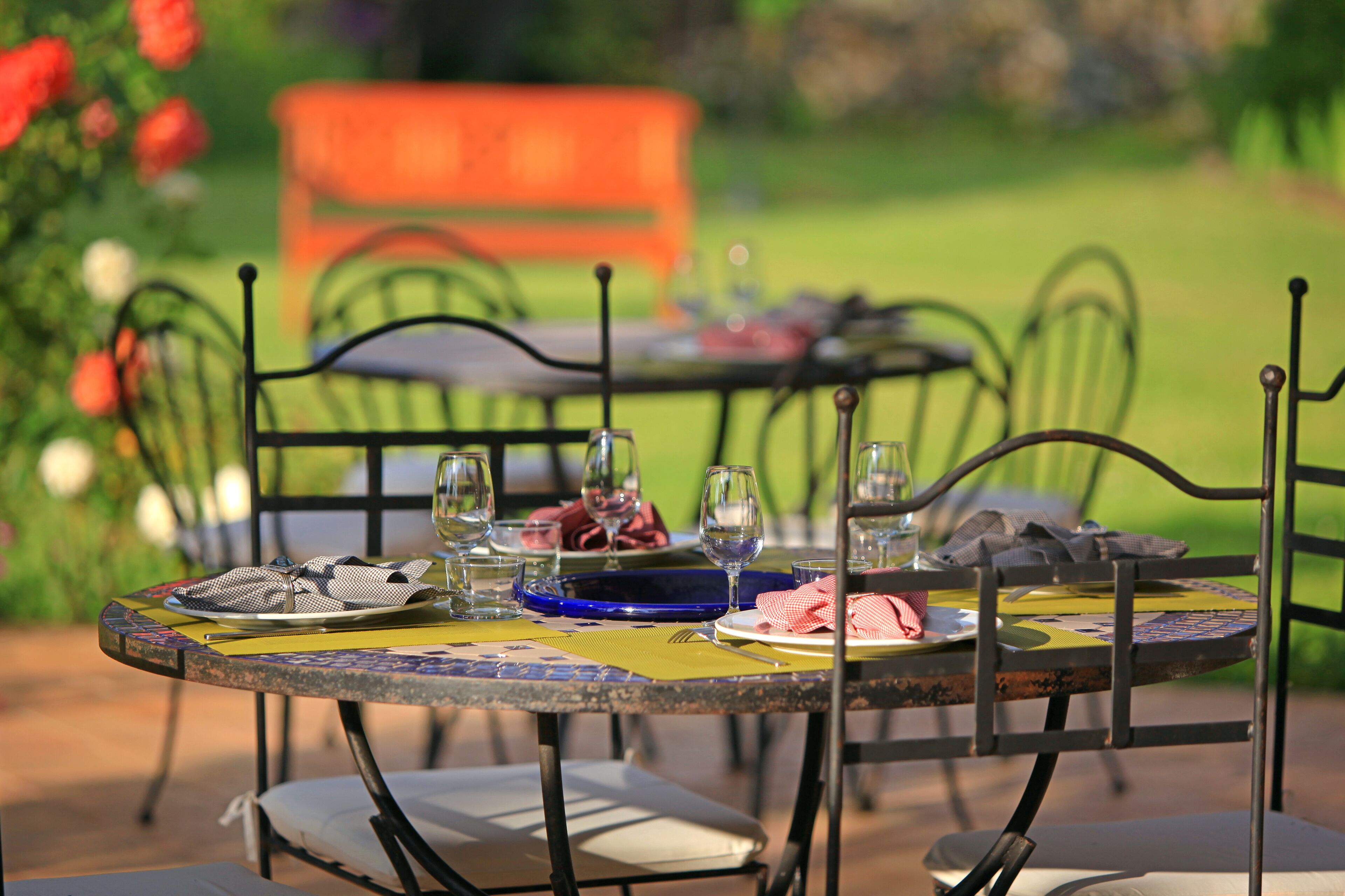 A round outdoor table with metal chairs, set with plates, glasses, and cutlery.