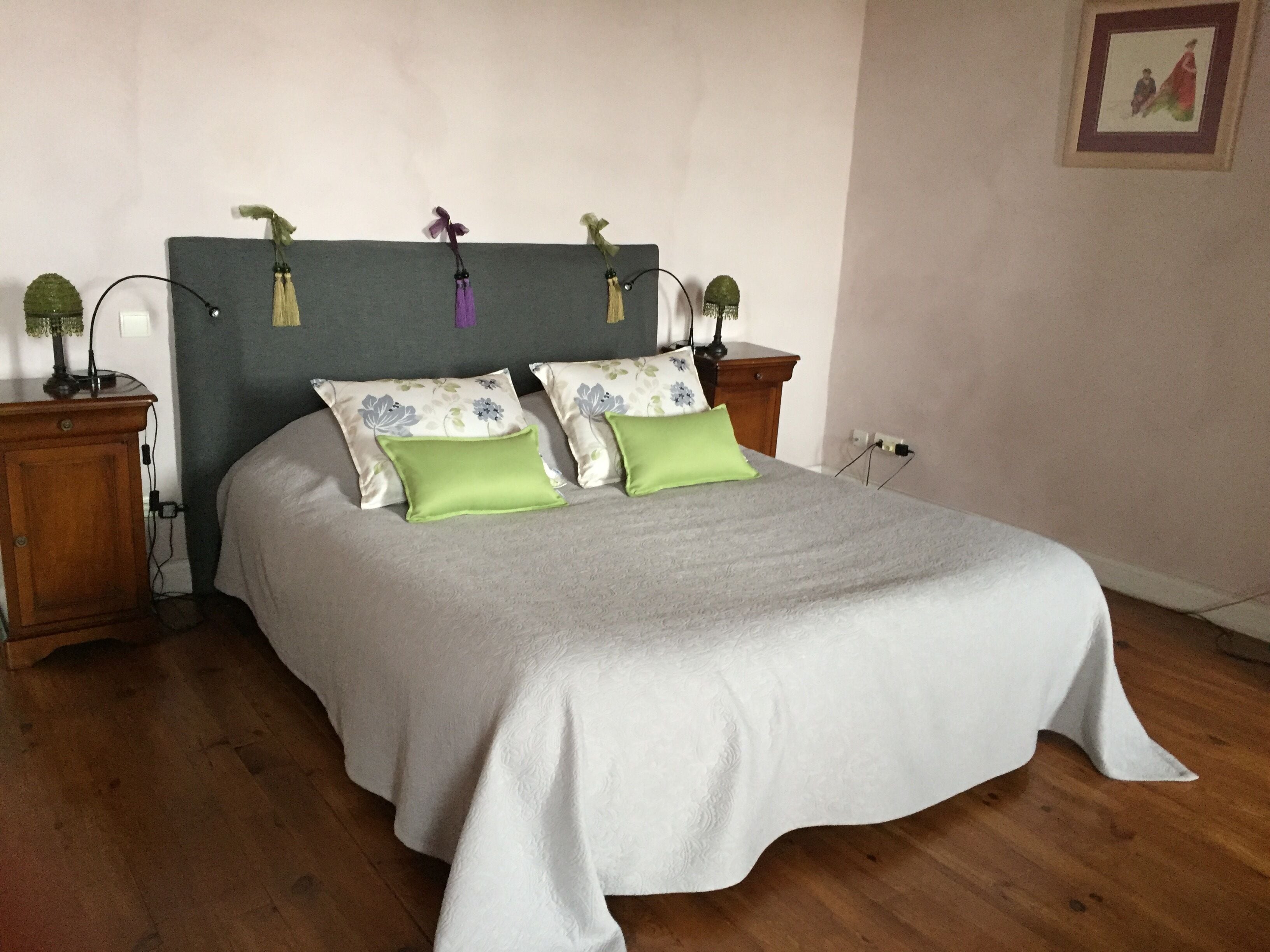 A neatly made bed with a grey bedspread and two pillows, one green and one blue with floral patterns. There are two bedside tables with lamps, and a framed picture on the wall.