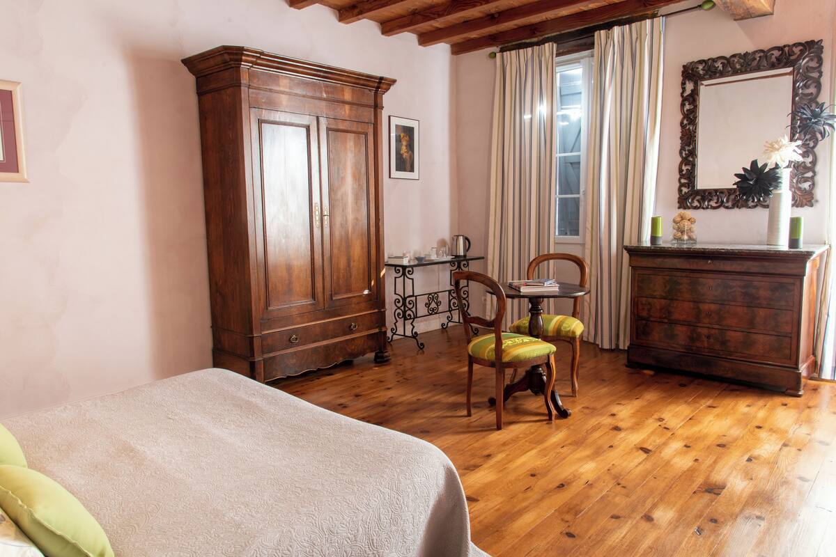 Un dormitorio con suelo de madera, una cama con colcha blanca, un armario de madera, una silla con cojín verde, una mesita de noche con lámpara y un espejo en la pared.