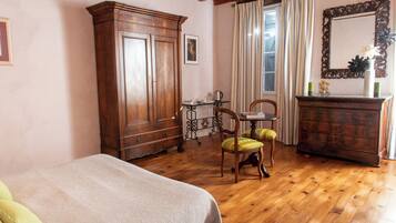 Un dormitorio con suelo de madera, una cama con colcha blanca, un armario de madera, una silla con cojín verde, una mesita de noche con lámpara y un espejo en la pared.