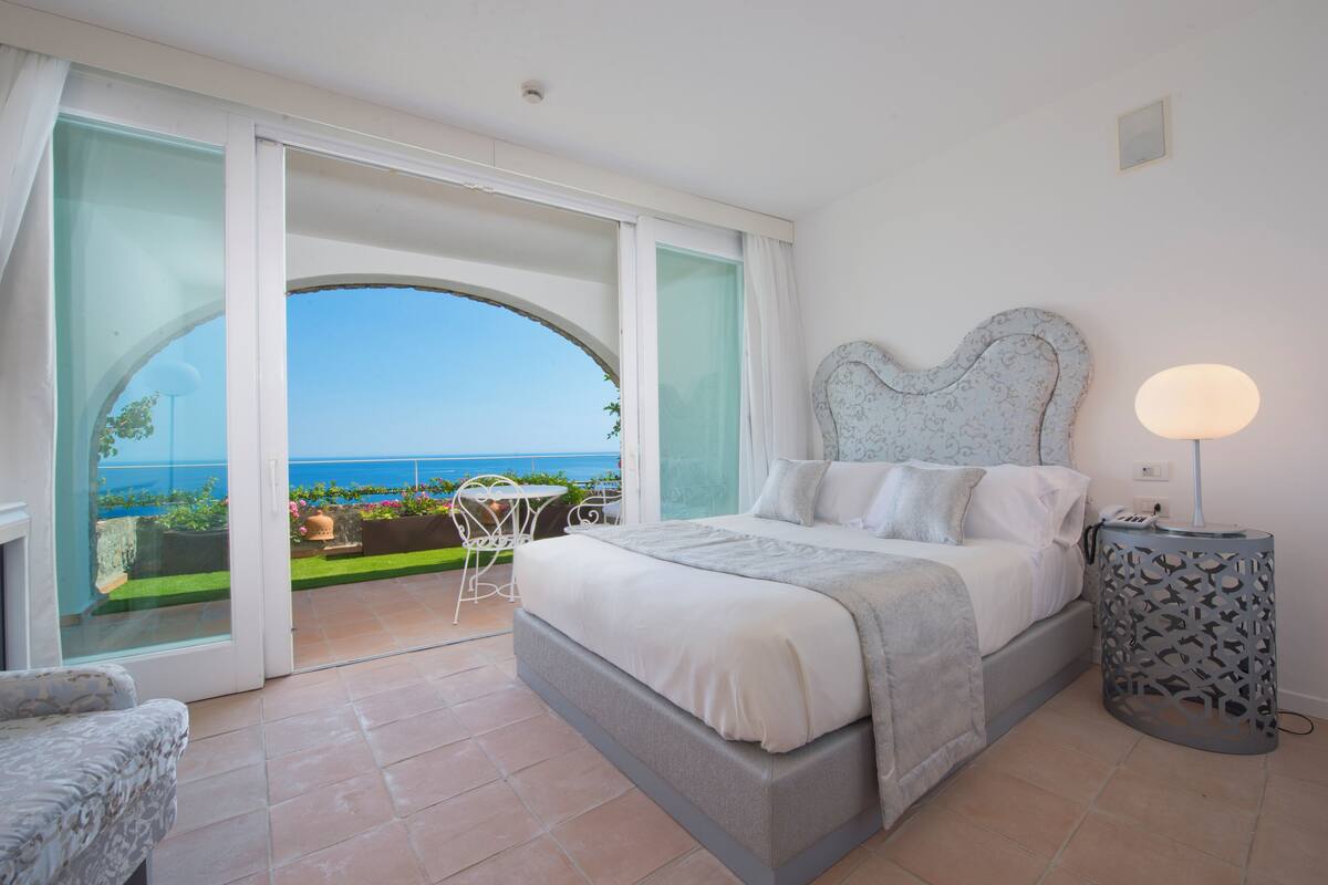 A bedroom with a large bed, a bedside lamp, and a view of the sea through a glass door.