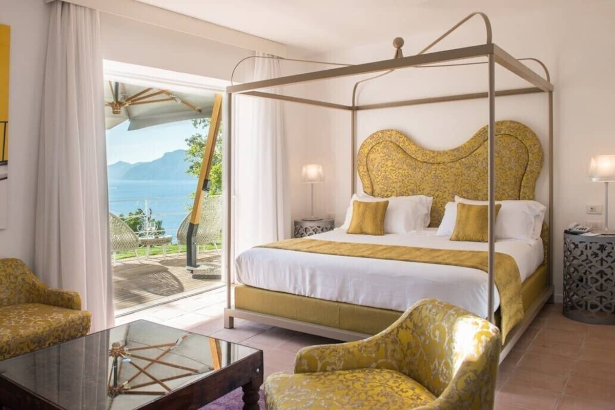 A four-poster bed with a gold headboard, white bedding, and yellow accents. There is a glass table with a decorative centerpiece, a yellow armchair, and a view of the sea through large windows.