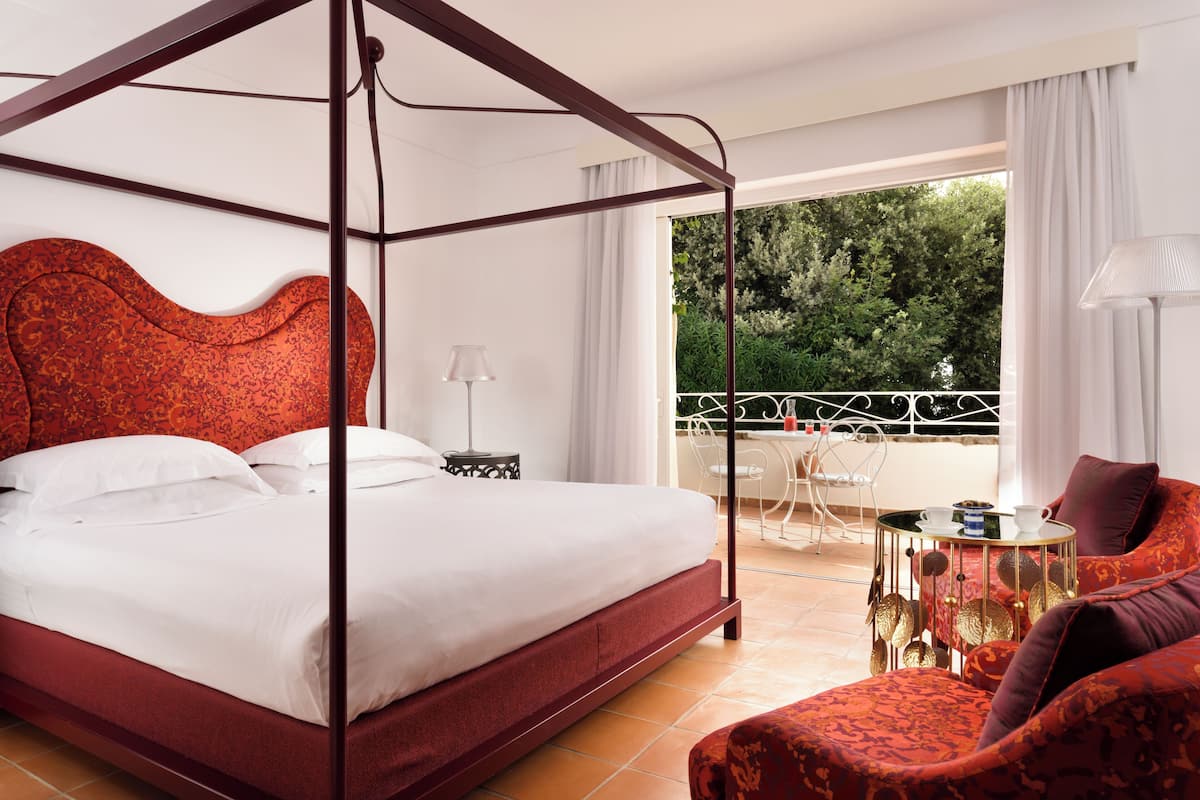 A four-poster bed with a red headboard, a balcony with a table and chairs, and a view of greenery outside.