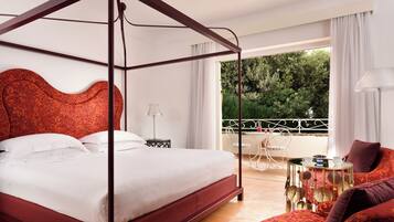 Una cama con dosel y cabecera roja, un balcón con mesa y sillas, y vistas al verde exterior.