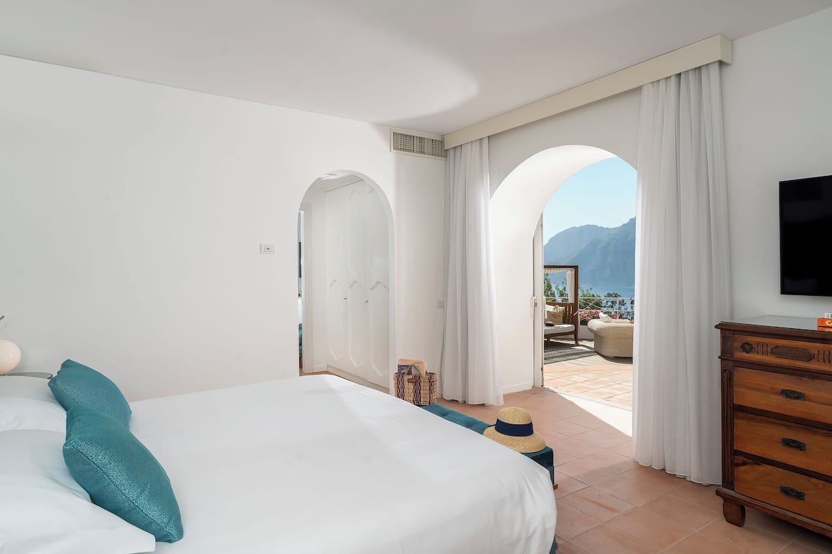 A bedroom with a large bed, a wooden dresser, and a TV. There is a view of mountains through an open door.
