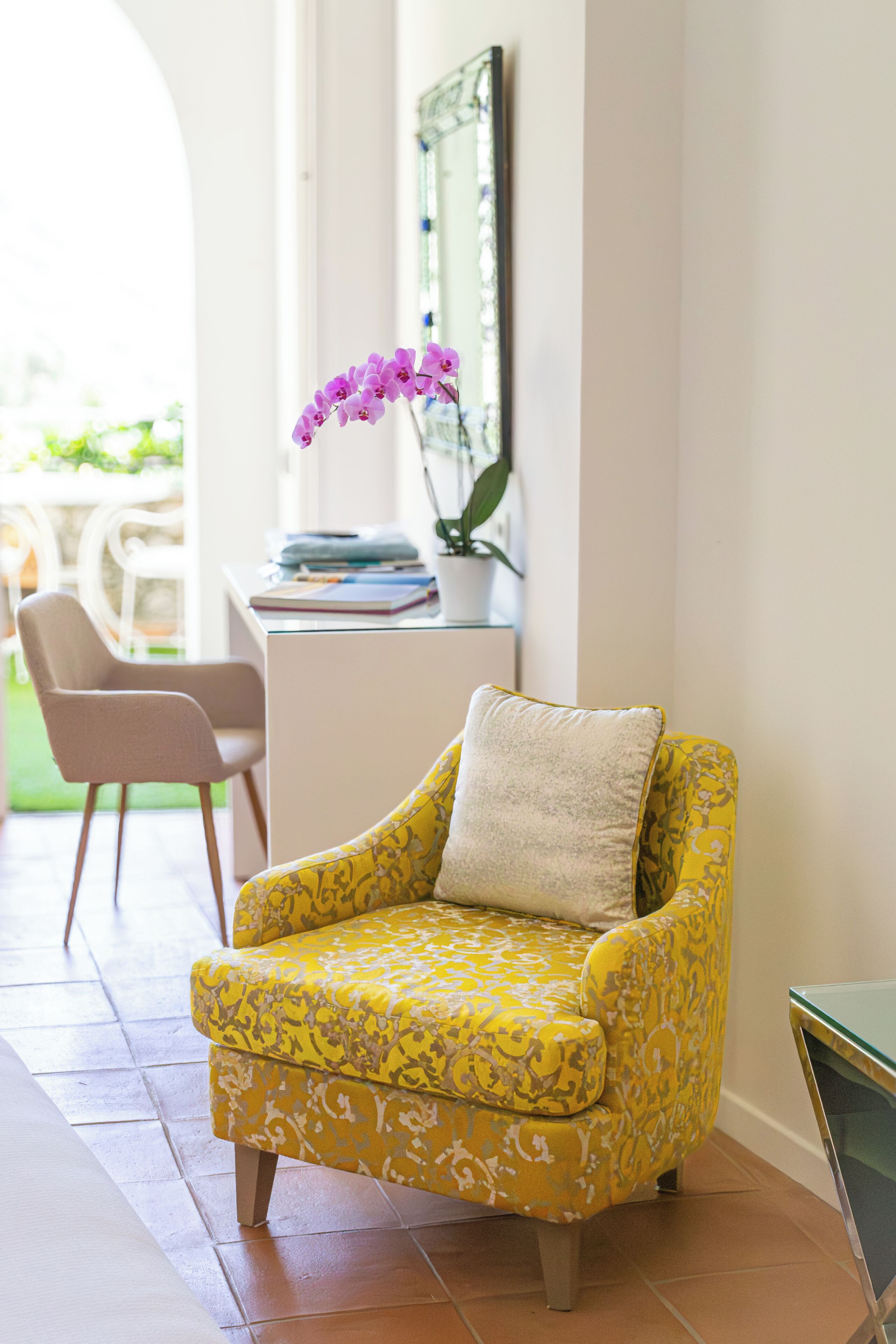 A yellow armchair with a cushion, a small table with a vase of flowers, and a chair in a bright room with a tiled floor.