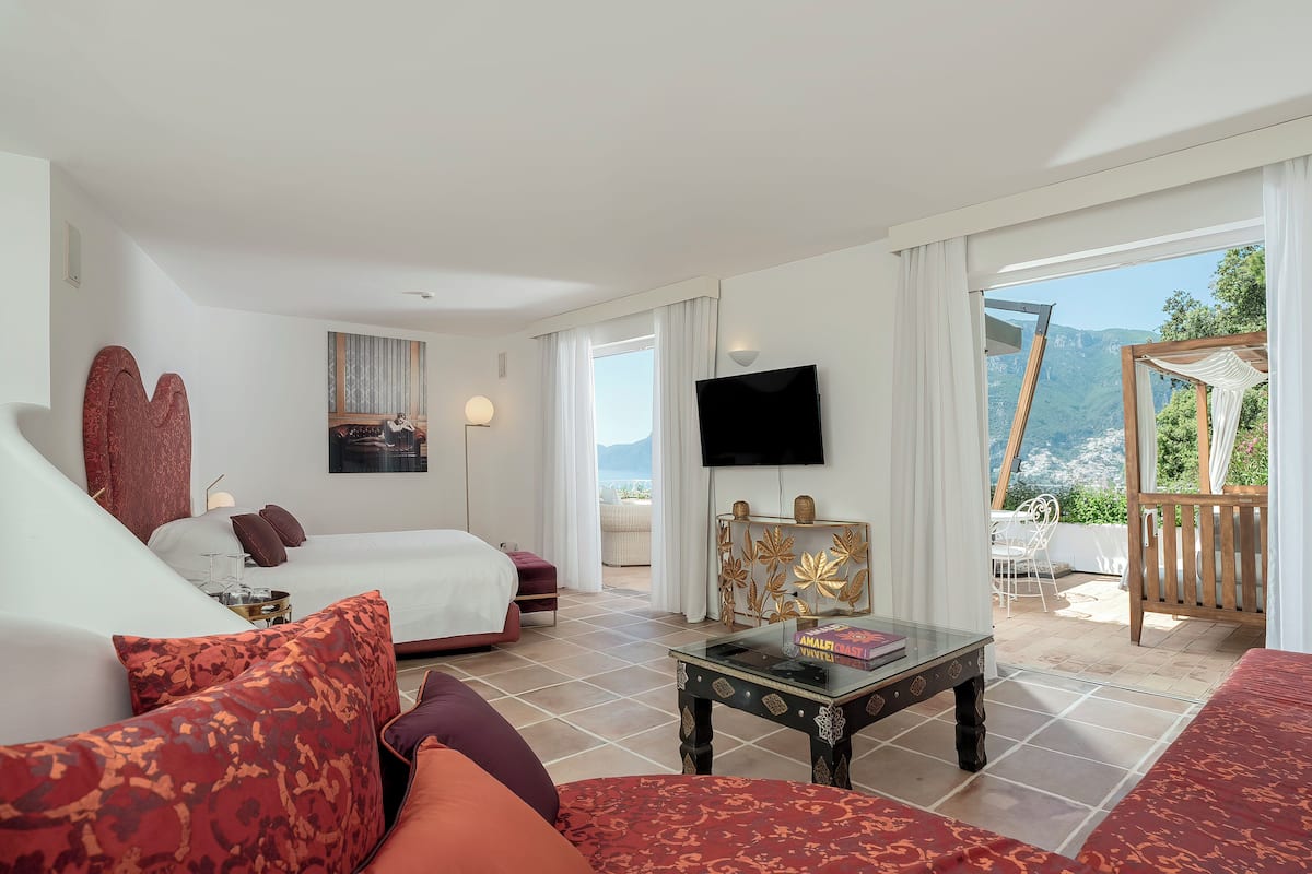 A hotel room with a bed, a sofa, a TV, and a balcony with a view of the mountains.