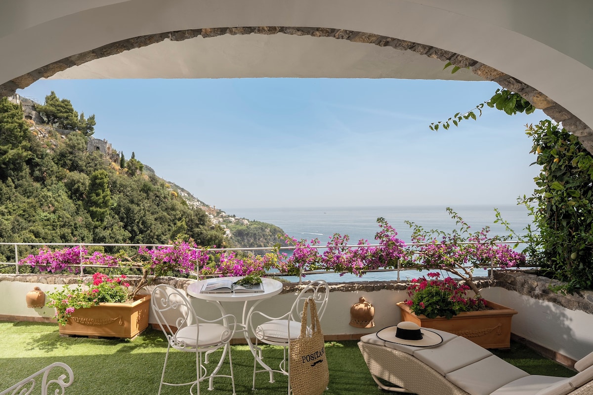 Une terrasse avec une table et des chaises, entourée de plantes et de fleurs en pot, d’où l’on peut admirer un paysage côtier.